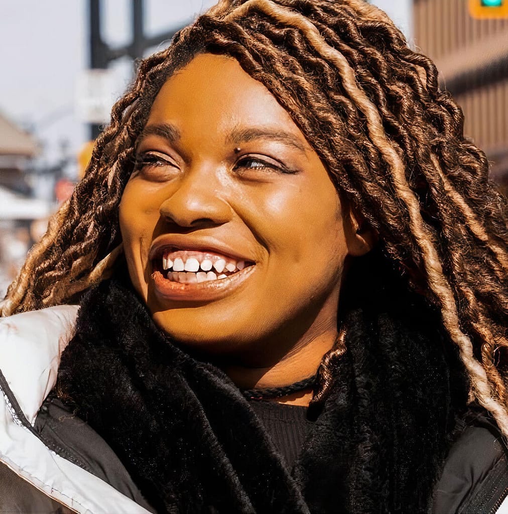 Headshot of Ciara Diane, a person with dark skin and light brown, curly, and textured hair with blonde highlights. They are smiling brightly, looking off to the side, and appear to be outdoors in a city setting. The person is shown from the chest up, dressed warmly in a black scarf and a light-colored jacket, with sunlight shining on their face, highlighting their joyful expression. The blurred background suggests an urban environment, with buildings and traffic signals visible.