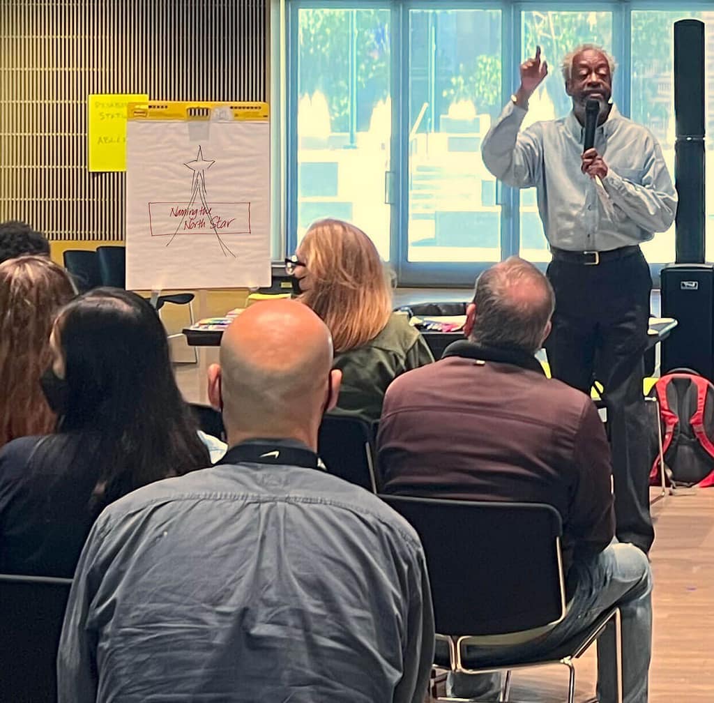 Harry McCord, a dark-skinned man with gray hair wearing a blue dress shirt, stands at the front of a room, holding a microphone and speaking to an audience. He gestures upward with one hand, emphasizing a point. Behind him is a flip chart with a drawing of a star and the words ‘Naming Our North Star.’ The audience, consisting of people with light and dark skin tones, is seated and focused on the speaker. The setting appears to be a seminar or workshop, with sunlight streaming through the windows in the background.