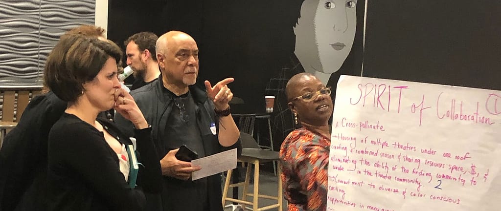 A group of people engaged in a discussion around a large poster labeled ‘SPIRIT of Collaboration.’ A person with dark brown skin and short hair, wearing glasses and a patterned outfit, is pointing to the text on the poster. Another individual with light skin and short hair, appearing to be deep in thought, stands nearby with their hand on their chin. A man with light skin and a bald head, wearing a dark shirt, gestures while holding a notepad, actively participating in the conversation. The background features a modern interior with a wall design and abstract artwork, suggesting the setting is likely a creative or collaborative space.