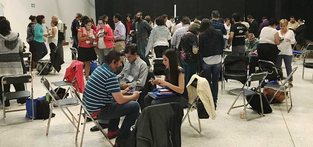 A large group of people in a spacious indoor setting, engaged in discussions and activities. Some are seated in pairs or small groups on folding chairs, while others stand and interact. The atmosphere suggests a workshop, networking event, or training session, with many participants taking notes, using tablets, or conversing.