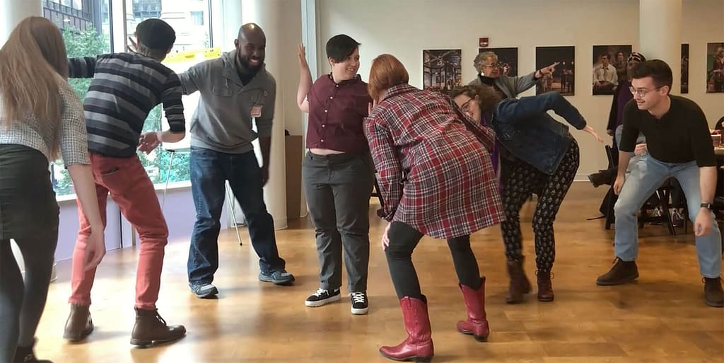 A group of people with diverse skin tones are engaged in a lively, interactive activity, bending, and moving in different directions in a group exercise led by Keryl McCord who stands at the back of the room pointing off to the right. The participants are in a brightly lit room with wooden floors and large windows, allowing natural light to fill the space. The atmosphere is playful and energetic, with everyone actively involved and seemingly enjoying the moment. One person in the foreground is wearing red boots, adding a pop of color to the scene.