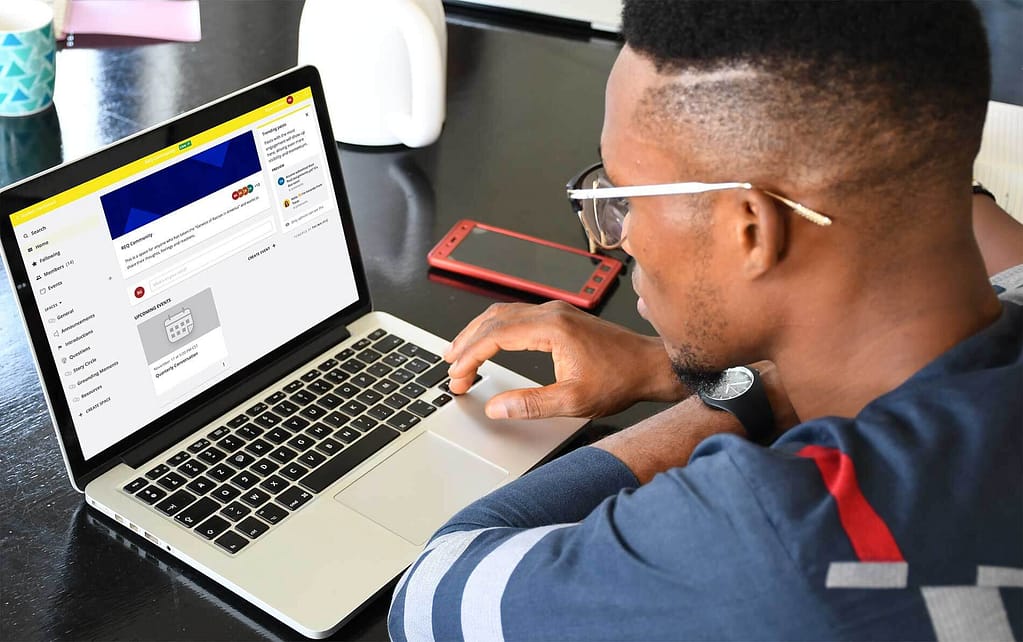 This image shows a Black man using a laptop, looking at what appears to be an online community platform. The screen displays an online learning platform with various community interactions. A red smartphone is placed next to him on the table, and he is concentrating on his computer work, likely engaged with the platform's features. The setting suggests a casual work-from-home or remote-learning environment.