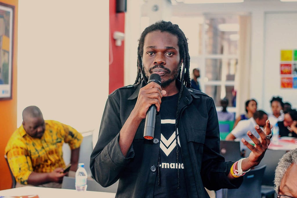 A man with dark skin and long dreadlocks holds a microphone and speaks while gesturing with his other hand. He is dressed in a black jacket and stands in a room with other people seated in the background. The atmosphere suggests a discussion or presentation, with the man appearing focused and engaged with his audience. A person with light brown skin wearing a yellow patterned shirt is visible in the background, looking at something on his phone.