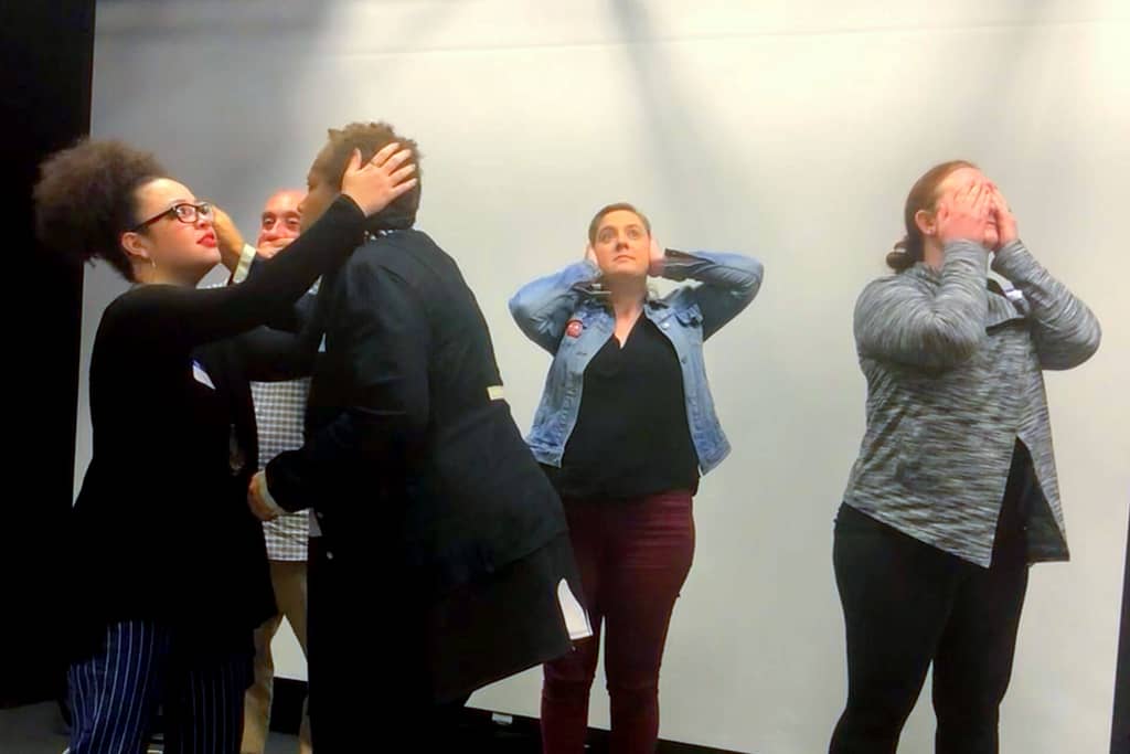 A group of five people with diverse skin tones are standing in a line, each engaging in a different action or gesture. A woman with light brown skin and curly hair, wearing glasses, is gently holding the face of another person who appears to be listening intently. Behind them, a light-skinned woman with short hair is holding her hands over her ears, while another light-skinned woman covers her face with her hands. A third light-skinned man, barely visible between the front two women, has his hand over his mouth. The setting appears to be a theatrical or expressive workshop, where participants are exploring different emotions or communication styles through physical gestures. The atmosphere is one of focused engagement and interaction.