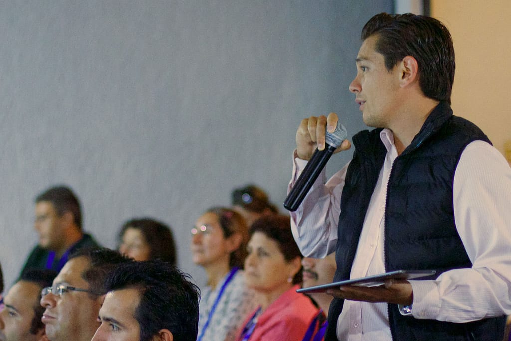 A light-skinned man is standing and speaking into a microphone while holding a tablet in his other hand. He is wearing a white shirt and a black puffer vest, addressing an audience that is seated and listening attentively. The setting appears to be a conference or seminar, with several people visible in the background, all focused on the speaker. The atmosphere suggests a formal or professional discussion.