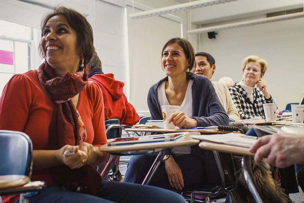 This image shows a group of people seated at desks in a classroom setting, appearing to be engaged in a lecture or discussion. A woman in the foreground, with medium skin tone and wearing a red top and a scarf, smiles while looking toward the front of the room. To her right, another woman with a light skin tone, wearing a white shirt and a blue cardigan, holds a cup and also smiles as she listens. The environment suggests a positive and interactive learning atmosphere, with a diverse group of participants seated at desks with notepads, laptops, and coffee cups.