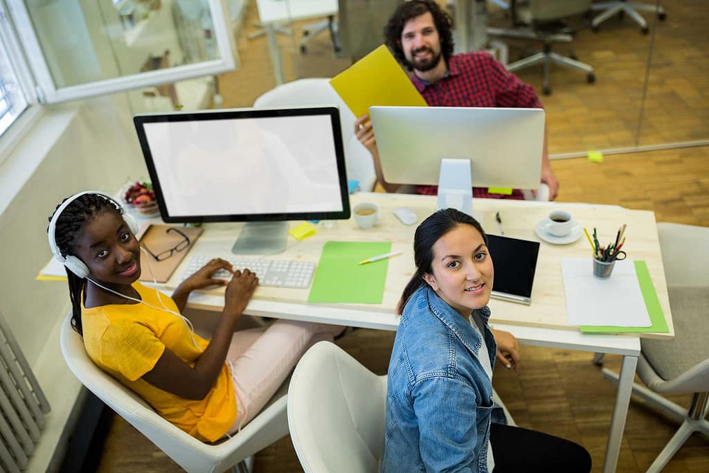 Three people with diverse skin tones are working together in a modern office space. A dark-skinned woman with braided hair is seated at a desk, wearing headphones and looking up from her work on a computer. A light-skinned woman with long dark hair is seated beside her, smiling at the camera, with a tablet and a cup of coffee on the desk. A light-skinned man with curly hair and a beard sits behind them, holding a yellow folder and smiling. The office is bright and organized, with additional desks and chairs visible in the background. The atmosphere appears collaborative and productive.