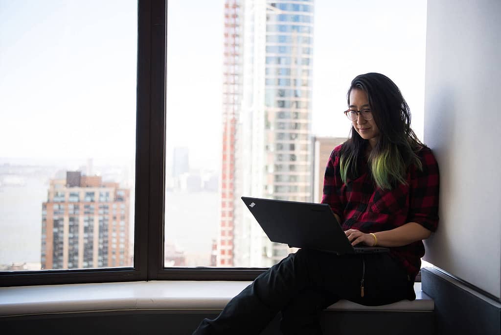 The image shows a woman sitting by a large window, working on a laptop. She has long black hair with green tips and wears glasses and a red and black checkered shirt. Outside the window, tall buildings and a distant city skyline are visible, suggesting she is in a high-rise building. The overall mood is calm and focused, with natural daylight illuminating the space.