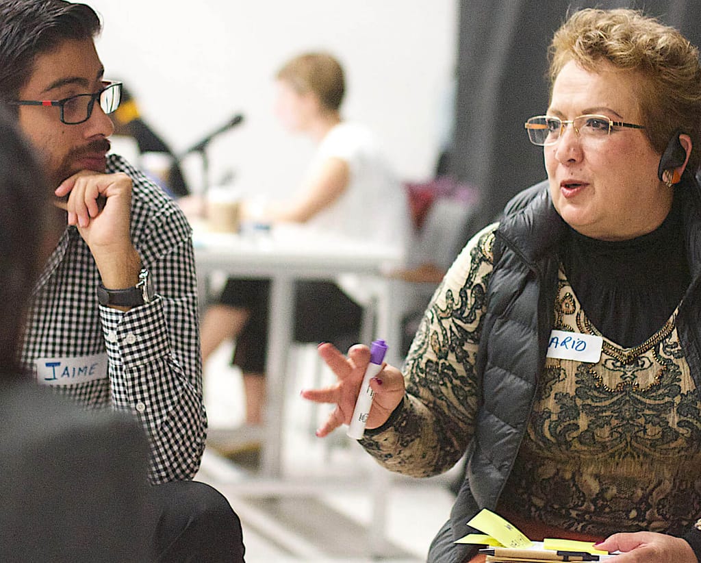 A light-skinned woman with short blonde hair, glasses, and a headset speaks while holding a marker and a stack of sticky notes. She is engaged in a conversation with a light-skinned man wearing glasses and a checkered shirt. Both have name tags, with the woman's reading ‘MARIA’ and the man's reading ‘JAIME.’ They appear to be in a group discussion or brainstorming session, with a blurred background of other participants working in the same space.