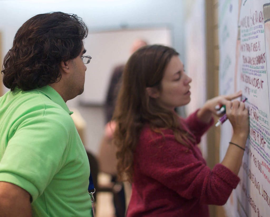 A light-skinned woman with long brown hair writes on a large sheet of paper on the wall, using colorful markers. Beside her stands a light-brown-skinned man with wavy dark hair, wearing glasses and a bright green polo shirt, who is attentively observing her work. The scene appears to be part of a collaborative brainstorming or planning session, with both participants focused on capturing ideas or information on the board.