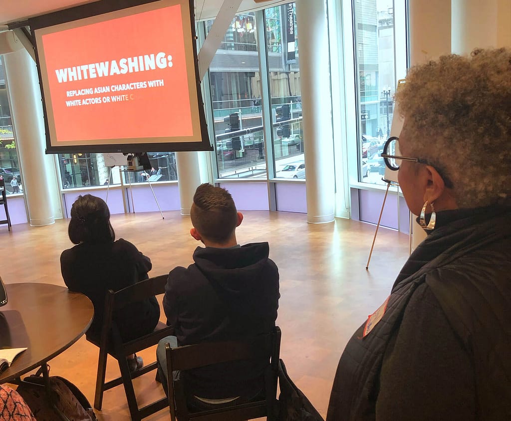 A group of people seated in a room with large windows, watching a presentation projected on a screen. The slide displayed reads, ‘WHITEWASHING: Replacing Asian characters with white actors or white…’ The person in the foreground, identified as Keryl McCord, has medium brown skin and short gray curly hair. She is wearing glasses and hoop earrings, standing and observing the presentation. Other attendees, with light skin and dark hair, are seated and facing the screen.