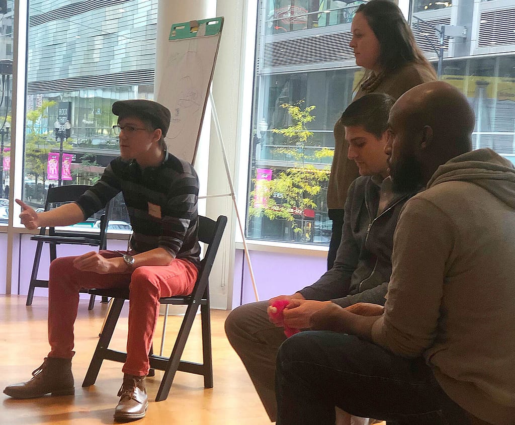 This image shows a group of four people engaged in a collaborative meeting or workshop. One person with light skin, wearing a striped shirt, red pants, and a cap, is actively speaking and gesturing while seated. To their left, a person with light skin stands, observing the conversation, while two others with medium brown skin and light skin are seated and listening attentively. The background reveals large windows showcasing an urban setting with buildings and street signs, suggesting a professional or educational environment with a casual yet focused atmosphere.