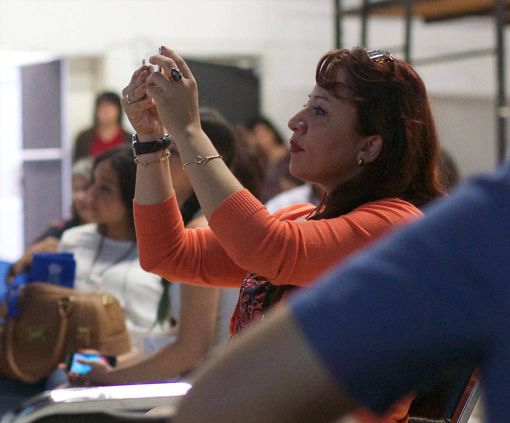 A woman with light brown skin and dark hair, wearing an orange sweater, taking a photo with her phone during an event. The individual is seated among other attendees, who are blurred in the background, indicating a focus on the act of capturing the moment. The woman is wearing jewelry, including bracelets and rings, and appears fully engaged in the activity. The setting suggests a meeting, presentation, or conference where participants are documenting the experience.