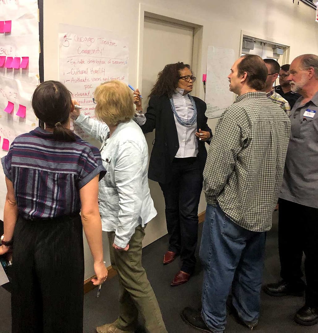A diverse group of people gathered around a large sheet of paper taped to a wall, engaged in a discussion. Several participants are writing on or pointing at the chart, which includes handwritten notes. The setting appears to be a workshop or community meeting, with sticky notes and additional charts on the wall, indicating a brainstorming or planning session.