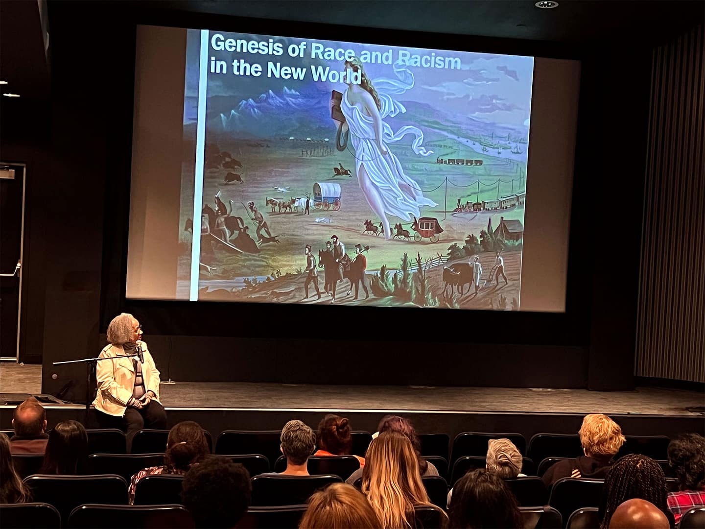 Keryl McCord sitting on a stage in front of an audience, giving a presentation. On the large screen behind her is a slide titled ‘Genesis of Race and Racism in the New World,’ featuring a historic painting of settlers traveling across a landscape with covered wagons and trains. Keryl, who has light gray hair, is seated with a microphone, and the audience is seated in rows of chairs, attentively watching the presentation. The setting appears to be a lecture hall or theater, and the overall atmosphere is academic and educational, focusing on historical themes related to race and racism.