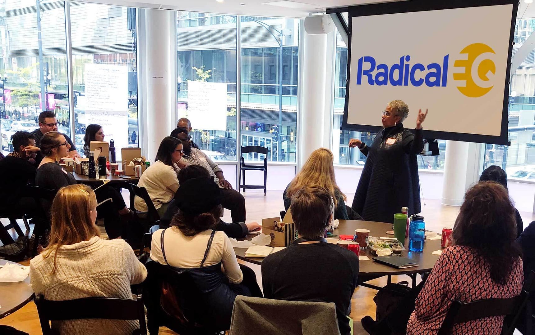 The image shows Keryl McCord giving a presentation to a seated audience in a modern, glass-walled room. The attendees are a diverse group, with a mix of light and dark skin tones, and are seated around tables, some of which have food containers and drinks. Keryl is standing in front of a projection screen displaying the words ‘Radical EQ’ in blue and yellow. The room has large windows with a view of a busy city street outside.