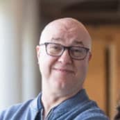 Headshot of Frank W. Spencer IV, a man with light skin, a bald head, and glasses, smiling warmly. They are wearing a blue sweater, and the background is softly blurred, suggesting an indoor setting with natural light coming through nearby windows. The individual’s expression is friendly and approachable, giving the image a relaxed and casual tone.
