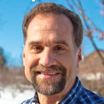 Headshot of David Brubaker, a man with light skin, short brown hair, and a beard with a mustache, smiling warmly at the camera. They are wearing a blue plaid shirt, and the background suggests an outdoor setting with a bright blue sky and some trees, possibly during winter as there is snow visible. The overall mood is cheerful and relaxed, with the person enjoying a sunny day outdoors.