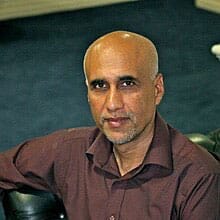 Headshot of Sohail Inayatullah, a man with medium brown skin and a bald head, dressed in a dark button-up shirt. They are seated indoors with a neutral expression, giving a calm and composed appearance. The background is dark, creating contrast and making the person stand out in the image. The overall tone suggests a professional or thoughtful atmosphere.