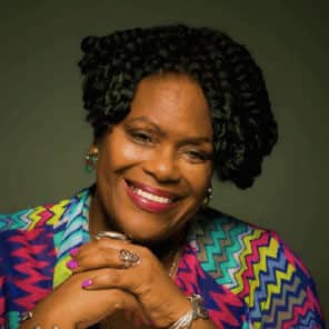 Headshot of Carol Bebelle, brown-skinned woman smiling brightly with curly black hair, wearing a colorful zig-zag patterned top in vibrant shades of blue, purple, yellow, and green. She is shown from the chest up, resting her chin on her hands, displaying silver jewelry, including rings and a bracelet, while posing against a dark green background. The overall feel is joyful and confident.