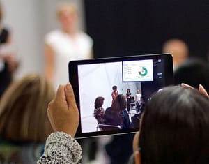 A person with light skin holds up a tablet, capturing a live event where a group of people are speaking in front of an audience. The tablet screen shows a pie chart and the speakers, who are blurred in the background of the image. The setting suggests a conference or presentation, with attendees seated and focused on the speakers. The image on the tablet provides a clearer view of the ongoing presentation.