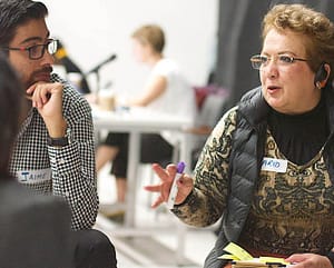 A light-skinned woman with short blonde hair, glasses, and a headset speaks while holding a marker and a stack of sticky notes. She is engaged in a conversation with a light-skinned man wearing glasses and a checkered shirt. Both have name tags, with the woman's reading ‘MARIA’ and the man's reading ‘JAIME.’ They appear to be in a group discussion or brainstorming session, with a blurred background of other participants working in the same space.