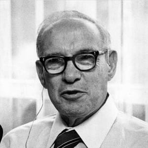 Black-and-white headshot of Peter Drucker, a notable figure in the field of management theory. He is seen wearing glasses and a formal shirt with a tie, with a background that appears to be softly lit, possibly by natural light through a window. His expression is neutral, and the photo captures him in a candid or conversational moment.