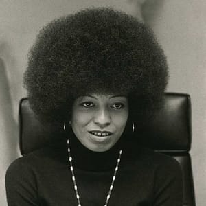 Black-and-white headshot of Angela Davis, a prominent political activist and scholar known for her work on civil rights, prison reform, and social justice. She is shown with an iconic Afro hairstyle, wearing a dark turtleneck and a long beaded necklace.