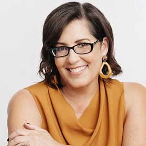 Headshot of Sue Dixon, a person with short, dark brown hair and glasses, smiling warmly. They are shown from the chest up, wearing a sleeveless mustard-yellow top and large geometric earrings. Their posture is relaxed, with arms gently crossed in front, and the background is a simple, light color, creating a professional yet approachable atmosphere. The person's overall expression is friendly and inviting.