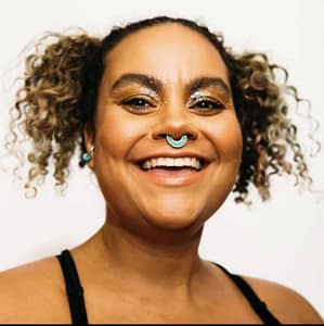 Headshot of adrienne maree brown, a woman with medium brown skin and curly hair styled in pigtails, smiling brightly. They have a septum piercing with a turquoise ring and are wearing bold, glittery eye makeup in gold tones. Their earrings are also turquoise, matching the septum ring. The person is dressed in a black top, and the plain white background emphasizes their joyful expression and vibrant style. The overall mood is playful and confident.