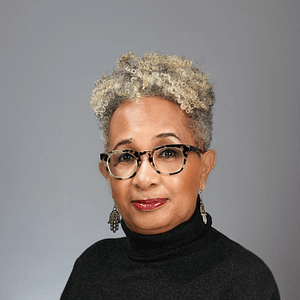 Headshot of Keryl McCord, a woman with medium brown skin and short, curly gray hair. She is shown from the chest up, wearing tortoiseshell glasses, a dark turtleneck, and dangling earrings. The expression on her face is calm and poised, with a neutral gray background that highlights her professional appearance.