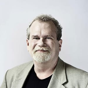 Headshot of Steve Scott, a man with light skin, short curly hair, and a light beard. He is shown from the chest up, wearing a beige blazer over a black shirt and is looking directly at the camera with a friendly expression. The background is a plain light color, giving the image a professional and clean appearance. His demeanor is approachable and calm.