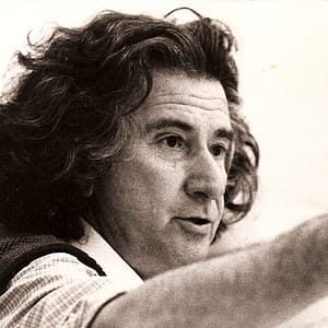 Black-and-white headshot of Augusto Boal, a person with light skin and thick, wavy, shoulder-length hair. They appear to be speaking or gesturing, with one arm extended outward. The individual is wearing a collared shirt and has an engaged, focused expression, possibly in the middle of a discussion or presentation. The image has a candid, dynamic feel, capturing the subject mid-action.