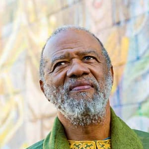 Headshot of John O’Neal, a man with dark skin and a gray beard, looking slightly upward with a thoughtful expression. They are wearing a green jacket over a patterned yellow top, and the background features a colorful, blurred mural or artwork. The lighting is soft, and the individual’s expression, combined with the vibrant background, gives the image a reflective and peaceful mood.