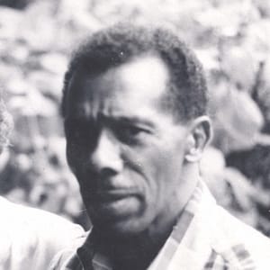 Black-and-white headshot of Jame Boggs, a man with dark skin and short, curly hair. The individual is wearing a checkered shirt and has a serious or contemplative expression. The background appears to be outdoors, with blurred foliage visible. The image has a vintage or historical feel, likely capturing a candid moment. The subject's expression and the overall tone of the photograph suggest a reflective mood.