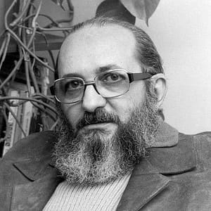 Black-and-white headshot of Paulo Freire, a man with light skin, wearing large, rectangular glasses, a full beard, and a mustache. They are dressed in a jacket over a sweater, with a calm and thoughtful expression. The background includes plants and possibly bookshelves, suggesting an intellectual or academic environment. The overall tone of the photograph is serious and reflective, capturing the individual in a relaxed yet contemplative pose.