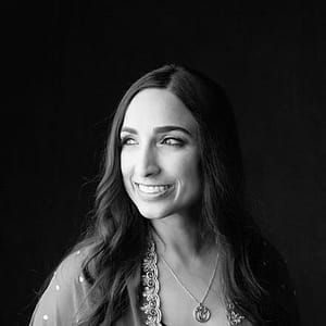Black-and-white headshot of Valarie Kaur, a woman with light skin and long, dark hair. They are smiling and looking off to the side. The individual is wearing a necklace and a patterned top or jacket with decorative trim. The lighting highlights their face, giving the image a warm and soft feel despite the monochrome palette. The overall tone is elegant and serene, capturing a candid, joyful moment.