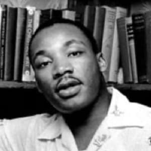 Black-and-white photo of Martin Luther King Jr., a man with dark skin and short hair, wearing a collared shirt. The individual is sitting or standing in front of a bookshelf filled with books, and they appear to be speaking or in the middle of a conversation, with a calm and thoughtful expression. The setting suggests an intellectual or reflective moment, with the books adding context to the environment.
