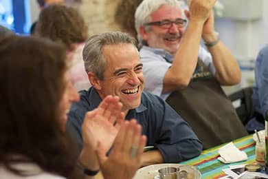 A group of people are seated at a table, sharing a moment of joy and laughter. The focus is on a man with gray hair who is smiling broadly, while others around him clap and laugh, creating a lively and celebratory atmosphere. The table is covered with a colorful striped tablecloth, adding to the festive mood.