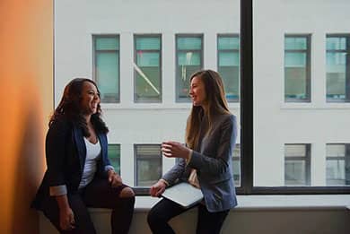 Two women sit on a windowsill in an office, engaged in a friendly conversation. Both are smiling, with one holding a laptop. Sunlight streams in from the left, creating a warm atmosphere, with the exterior of another building visible through the large window behind them.