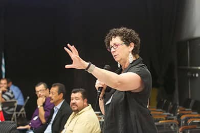 A woman with short curly hair and glasses speaks into a microphone while gesturing with her hand. She is addressing a group of seated men, who are attentively listening to her. The setting appears to be a conference or seminar with a dark background.