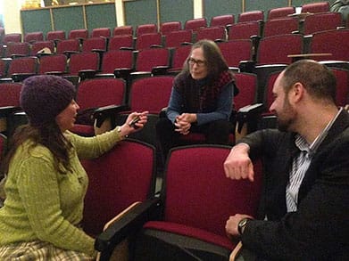 Three people with light skin are seated in a mostly empty auditorium with red chairs, engaged in a conversation. One woman, wearing a purple knit hat and green sweater, holds an object, possibly a recorder or phone, while speaking to a man in a suit and another woman with long gray hair and glasses, who is leaning forward attentively. The atmosphere appears casual and focused.