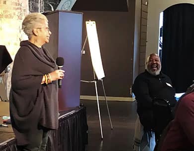 Keryl McCord, a woman with short gray hair, speaks into a microphone while smiling, addressing a man seated nearby who is laughing heartily. The setting appears to be a casual or semi-formal event, with a flip chart stand visible in the background and exposed brick walls adding to the atmosphere. The interaction between the two suggests a moment of humor or shared enjoyment.