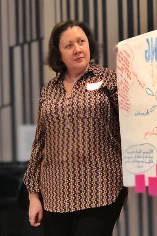 A light-skinned woman with dark brown hair is standing and holding up a large sheet of paper filled with handwritten notes and diagrams. She is wearing a patterned blouse and has a name tag on her chest. The setting appears to be a workshop or presentation, with the woman likely explaining or discussing the content on the paper. Her expression is focused and attentive.