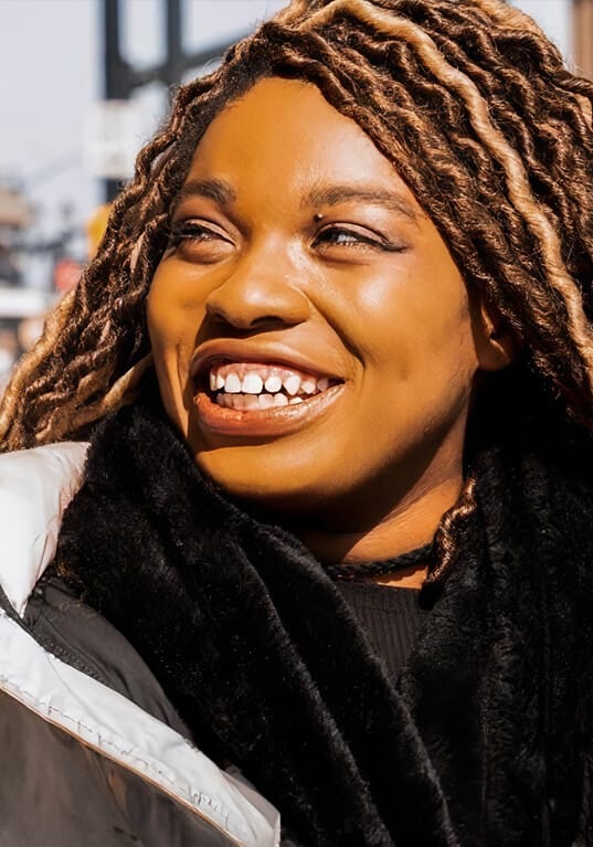 Headshot of Ciara Diane, a person with dark skin and light brown, curly, and textured hair with blonde highlights. They are smiling brightly, looking off to the side, and appear to be outdoors in a city setting. The person is shown from the chest up, dressed warmly in a black scarf and a light-colored jacket, with sunlight shining on their face, highlighting their joyful expression. The blurred background suggests an urban environment, with buildings and traffic signals visible.