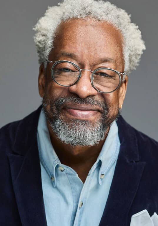 Headshot of Harry McCord, a brown-skinned man with gray curly hair and a beard is wearing round glasses and a friendly smile. He is shown from the chest up, dressed in a light blue denim shirt under a dark blue velvet blazer, with a white pocket square neatly folded in his jacket pocket. The background is a neutral gray, which draws attention to his warm expression and distinguished appearance. The image is a well-composed portrait that highlights his personality and style.