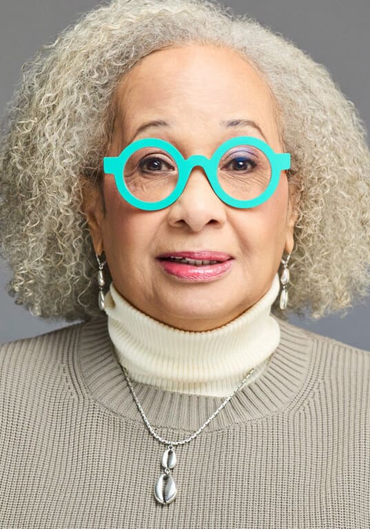 Headshot of Keryl McCord, a brown-skinned woman with gray curly hair wearing bright turquoise glasses, a beige sleeveless sweater over a cream turtleneck, and a silver necklace with a matching pendant. She is shown from the chest up, smiling slightly and looking directly at the camera, with a neutral gray background behind her. The image is a well-lit portrait that highlights her distinctive style and warm expression.