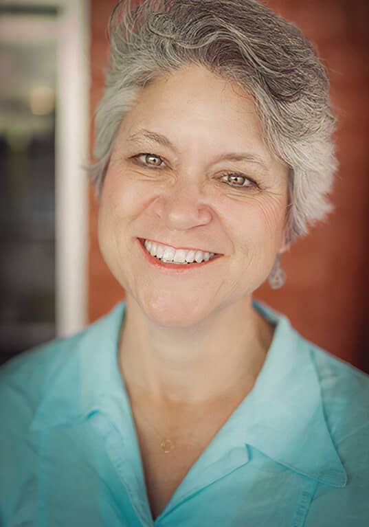 Headshot of Lisa Mount, a light-skinned woman with short gray hair, smiles warmly at the camera. She is shown from the chest up, wearing a light blue blouse, and her expression exudes friendliness and approachability. The background is softly blurred, featuring warm tones, which keeps the focus on her face. The overall composition suggests a candid and welcoming portrait.