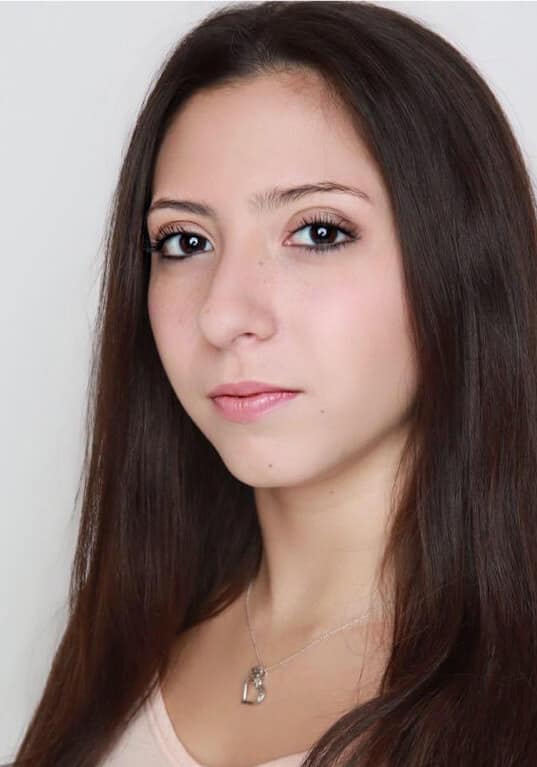 Headshot of Nisa Betancourt, Ta person with long, dark brown hair, wearing minimal makeup and a neutral expression. They are shown from the chest up, have light skin, dark eyes, and are dressed in a light-colored top. A delicate necklace with a small pendant is visible around their neck. The background is plain white, giving the image a clean and professional feel. The individual is looking directly at the camera with a calm and composed demeanor.