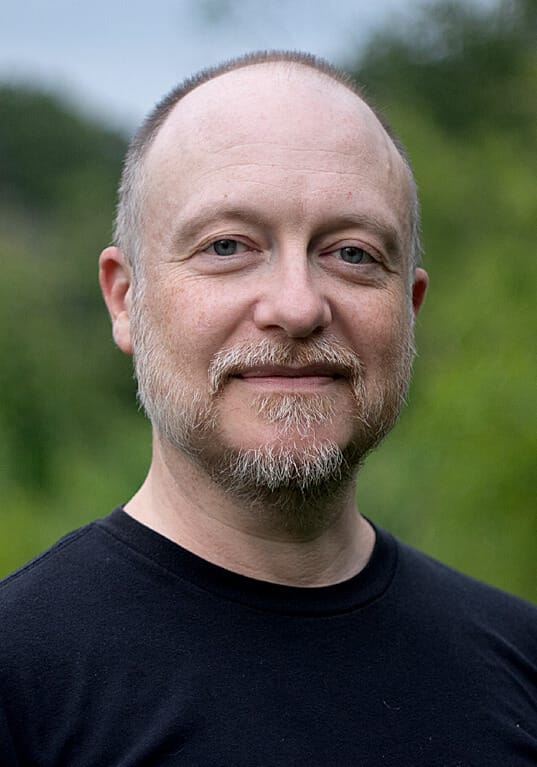 Headshot of Brooks Emanuel, a light-skinned man with a trimmed beard and short, thinning hair. He is shown from the chest up, wearing a black shirt and smiling slightly, with a blurred natural background of greenery. The soft lighting adds a calm and approachable feel to the portrait.